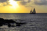 A single sailboat silhouetted against a golden sunset on a calm sea near rocky shores. Aruba, The Netherlands [IBR125026135]