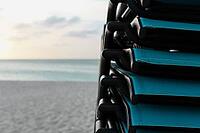 Stacked turquoise beach chairs facing a tranquil ocean and sandy beach during sunset [IBR125026143]