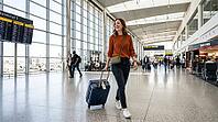 A smiling woman pulls her suitcase through a bright, modern airport concourse, with departure boards and shops visible, AI generated [IBR125026408]