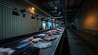 Industrial textile sorting facility conveyor belt with blurred mixed donated clothing in motion, NIR sensor and camera arrays mounted overhead on steel rail, automated fiber identification and recycling concept, no people, studio shot, AI generated [IBR125167443]