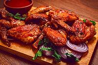 Cooked chicken wings lay on a wooden board. There are slices of onion and cilantro around the wings. A bowl of red sauce is next to the wings for dipping [IBR125177929]