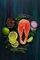 A salmon fillet is placed on a black stone board. Surrounding it are slices of lemon and lime, red onion, herbs, and a bowl of mixed pepper. The setting is on a dark wooden table [IBR125177930]