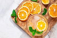 Slices of orange and lemon are arranged on a wooden board next to a glass of drink. Fresh mint leaves add color. The setting looks bright and inviting [IBR125177941]