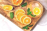 Slices of orange and lemon are arranged on a wooden board next to a glass of drink. Fresh mint leaves add color. The setting looks bright and inviting [IBR125177942]