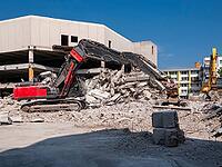 Red demolition excavator with grab in front of demolition building [IBR125180344]