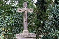A tall stone cross is placed in a wooded area. Green trees and bushes surround the cross. Small flowers grow at its base. The scene shows nature around the structure during the day, Borken, NRW, Germany [IBR125185853]