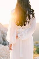 Bride is standing on a mountain touching her other hand with one hand in bright sunlight. Back view. [IBR125188163]