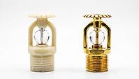 Two new fire sprinkler heads side by side, left with cream-white wax corrosion-resistant coating, right with standard bright brass finish, fire protection maintenance decision and chemical facility safety concept, white background, studio shot, AI generat [IBR125200731]