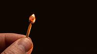 Single wooden matchstick in moment of ignition, tiny orange-white phosphorus flame just forming at tip, fingers lit from match-light below, Glimmers of Wonder winter hope and intimate festive light concept, dark background, studio shot, AI generated [IBR125200735]