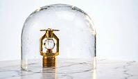 Brand-new brass fire sprinkler head displayed inside sealed clear glass bell jar dome with faint inner condensation, invisible corrosive atmosphere and chemical storage safety concept, white marble base, studio shot, AI generated [IBR125200738]