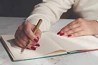 A person writes on an open notebook using a gold pen while sitting at a wooden table. The scene takes place in daylight. The focus is on the writing activity [IBR125204123]