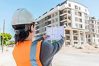 A young Hispanic woman in a hard hat and safety vest examines blueprints at a construction site with a partially built apartment building in the background [IBR125237468]