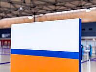A colorful sign with orange, white, and blue sections stands in an indoor space of airport or terminal. The background features a modern architectural design [IBR125237489]
