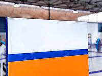 A colorful sign with orange, white, and blue sections stands in an indoor space of airport or terminal. The background features a modern architectural design [IBR125237495]