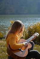 Blonde woman playing string guitar outdoors in autumn forest. Concept of sound therapy, mental health and wellness rituals. Calmness tranquility audio-sensory practices. Aura farming energy holistic health [IBR125261599]