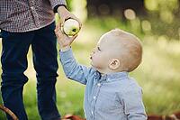 Crop of parent giving for little baby boy apple, walking together in summer park. Little boy in blue cap wearing in checked shirt smiling and having fun outside in nature. Summertime concept [IBR125261607]