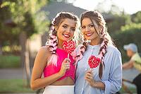 Pretty girlfriends with colorful braids holding candy heart on stick like heart and happy smiling at camera. Gorgeous and stylish women walking at street after shopping and having fun together [IBR125261614]