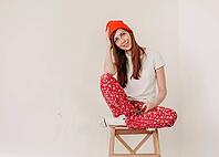 Smiling funny girl in a red hat and Christmas pajamas sitting in bed with white sheets. holidays for christmas. Good Christmas mood girl. Girl in a white T-shirt and pajamas, against the background of a white wall, copy space [IBR125261618]