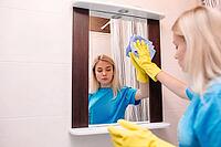 View of young blonde girl wearing in yellow protective gloves working at home, cleaning with spray and using squeegee to wash mirror at bathroom. Service and housekeeping concept [IBR125261623]