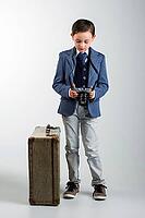 8 year old boy dressed up in suit, standing next to vintage suitcase, looking at photo camera taking up a vintage suitcase. travel concept [IBR125261637]