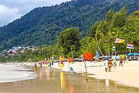 Kathu Phuket Thailand 08. October 2025 Patong Beach bay panorama view with promenade tourists people blue water waves sun loungers parasols lifeguard watch tower mountains and palm trees in Thailand [IBR125280359]