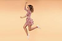 Side view of crazy girl wearing pink floral dress and white sneakers jumping and laughing in studio. Attractive young female in glasses enjoying warm weather and stylish outfit. Concept of happiness [IBR125324104]
