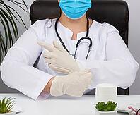 Woman doctor in a white uniform, disposable medical mask is sitting in a chair at the table and puts on his hands white sterile latex gloves, doctor?s office [IBR125331158]