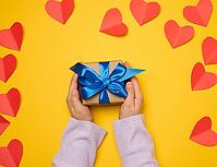 A woman's hand holds a gift box wrapped in a blue silk ribbon on a yellow background [IBR125331174]