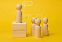 Wooden figures on a yellow background symbolize communication and leadership, one figure stands on a pedestal, representing a speaker conducting a dialogue with a team discussing ideas and solutions [IBR125344249]