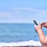A man holds a smartphone in his hands against the background of a blue sea. Black screen, layout. The concept of tourist travel and free time for people. With space to copy. [IBR125344261]