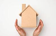 Female hands folded to each other over a wooden miniature model house on a white background. Real estate insurance concept, environmental protection, family happiness [IBR125344299]