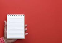 Female hand holding an open notebook with blank white pages on a red background, copy space [IBR125344301]