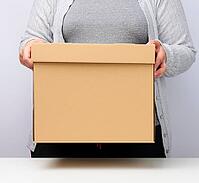 Woman in gray clothes stands and holds a brown box on a white background, moving, sending and delivering goods [IBR125344330]
