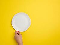 Female hand holding a white empty round plate on a yellow background, top view, copy space [IBR125363576]