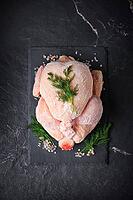 A top view of a whole frozen chicken on a black slate with dill and pink salt, vertical shot. The raw poultry meat, which is covered with frost, stands against a dark stone background [IBR125363613]