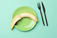 Round green ceramic plate with a whole banana and knife and fork on a green background, diet, top view [IBR125382567]