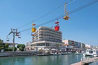 View of the Transcanal cable car with red and yellow gondolas and chairlift seats above the banks of the channeled Lez River in front of an apartment complex under a cloudless blue sky, Palavas-les-Flots, Herault Department, Occitania, France [IBR125405038]