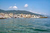View from the clear, moving water of the small bathing area on a gravel book in the harbor near the pier of the picturesque backdrop of the Old Port of Bastia with the church of Saint-Jean-Baptiste, the green lighthouse at the pier and a ferry docked in t [IBR125405049]