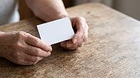 Older adult's wrinkled hands holding a small plain blank card above a modest worn wooden table, soft natural daylight, no face visible, vaccination record and preventive health concept, AI generated [IBR125407391]