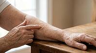 Close-up of an older adult's wrinkled hand pressing a round adhesive bandage onto the opposite forearm after vaccination, on a plain wooden table, soft natural daylight, no face visible, AI generated [IBR125407400]