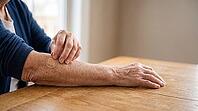 Close-up of an older adult's wrinkled hand pressing a round adhesive bandage onto the opposite forearm after vaccination, on a plain wooden table, soft natural daylight, no face visible, AI generated [IBR125407421]