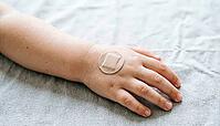 Close-up of a small child's hand and forearm with a plain round adhesive bandage after vaccination, on a soft grey fabric surface, gentle natural daylight, no face visible in frame, AI generated [IBR125407425]