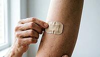 Adult hands applying a small plain beige nicotine patch to the opposite upper arm, fingers smoothing the edge, soft natural daylight, no face visible, smoking cessation health concept, AI generated [IBR125407426]