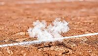 Fine white chalk dust suspended mid-air above a reddish-orange clay tennis court's boundary line, freshly disturbed mark below, bright daylight, decisive line call sports concept, AI generated [IBR125407427]