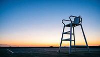 Empty white umpire chair silhouetted against a gradient dusk sky beside an unmarked tennis court, warm horizon light fading to deep blue, quiet reflective mood, sports atmosphere concept, AI generated [IBR125407440]