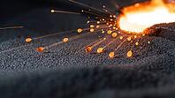 Tiny glowing orange metal spatter droplets flying from the edge of a molten weld pool above grey metal powder, frozen mid-flight, industrial macro photography, additive manufacturing, AI generated [IBR125407441]