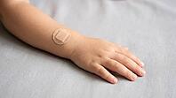 Close-up of a small child's hand and forearm with a plain round adhesive bandage after vaccination, on a soft grey fabric surface, gentle natural daylight, no face visible in frame, AI generated [IBR125407461]