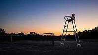 Empty white umpire chair silhouetted against a gradient dusk sky beside an unmarked tennis court, warm horizon light fading to deep blue, quiet reflective mood, sports atmosphere concept, AI generated [IBR125407472]