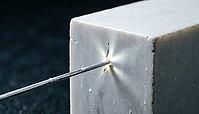 Thin silver-grey metallic lithium dendrite filament piercing through a fracture point in a solid crystalline ceramic block, charcoal background, studio lighting, battery failure science, AI generated [IBR125407474]