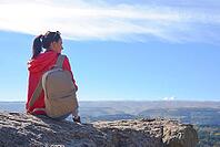 A tourist with a backpack is sitting on a mountain peak and enjoying the beautiful view. A wandering woman in an orange jacket is resting on a rock and looking out at the beautiful sunny landscape of the Caucasus [IBR125411136]